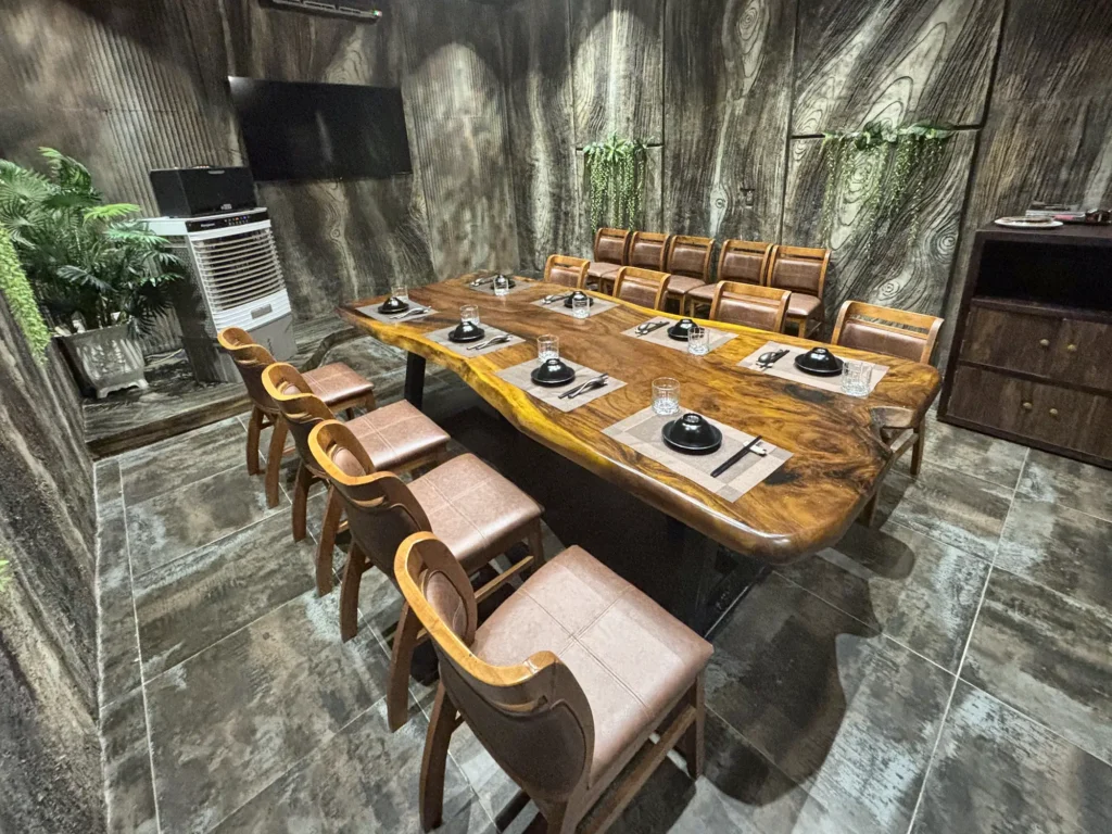 Phòng VIP 01 (Hồng Châu) tại Gánh Ngon: bàn gỗ dài, ghế bọc nệm nâu, tường giả vân gỗ xám và cây xanh, TV treo tường