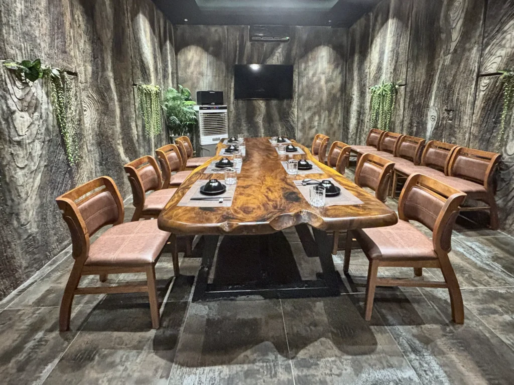 Phòng VIP 01 (Hồng Châu) tại Gánh Ngon: bàn gỗ dài, ghế bọc nệm nâu, tường giả vân gỗ xám và cây xanh, TV và quạt làm mát cuối phòng