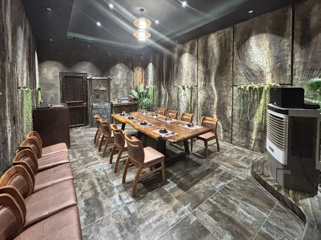 Phòng VIP 01 (Hồng Châu) tại Gánh Ngon: bàn gỗ dài, ghế bọc nệm nâu, tường giả vân gỗ xám và cây xanh — toàn cảnh phòng