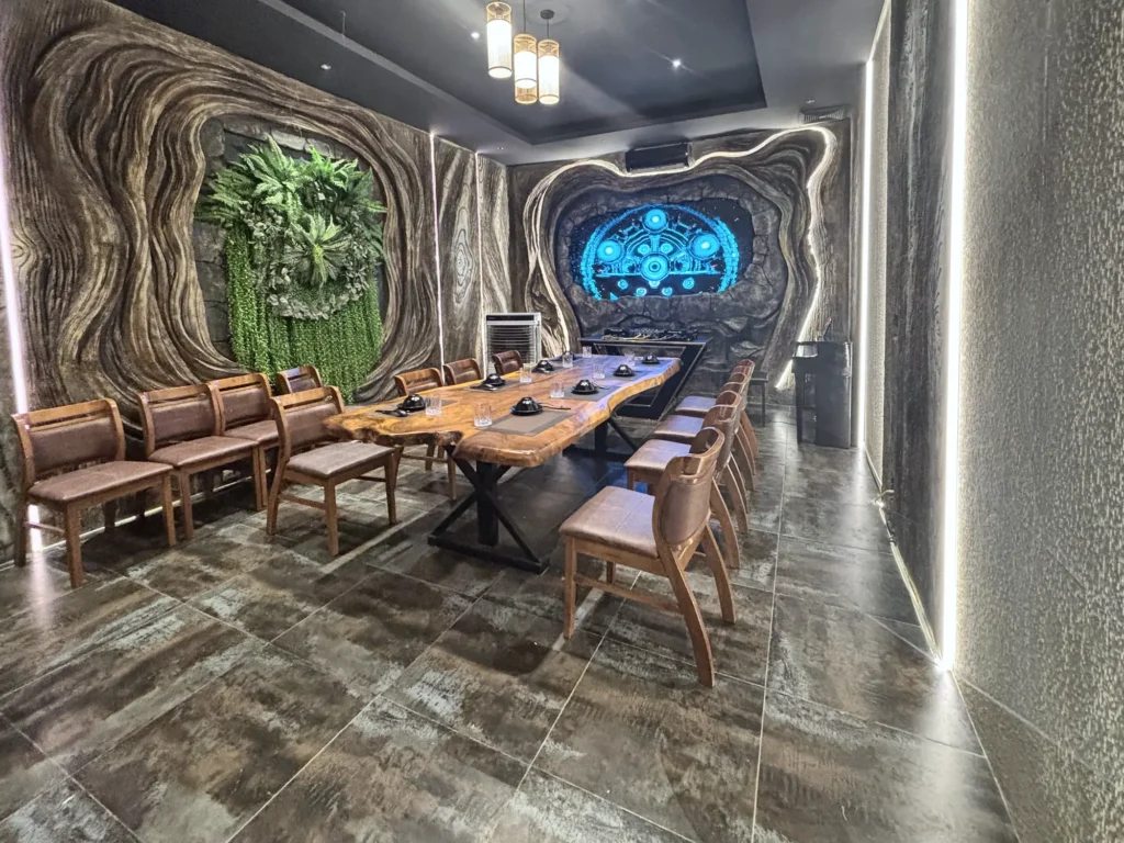 Phòng VIP 02 (Thiên Châu) tại Gánh Ngon: bàn gỗ dài, ghế bọc nệm nâu, tường vân gỗ uốn lượn, mảng cây xanh và đèn LED viền, màn hình trình chiếu cuối phòng