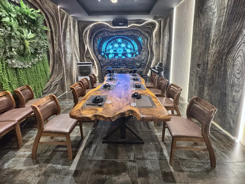 Phòng VIP 02 (Thiên Châu) tại Gánh Ngon: bàn gỗ dài, ghế bọc nệm nâu, tường vân gỗ uốn lượn, mảng cây xanh và đèn LED viền, màn hình trình chiếu cuối phòng