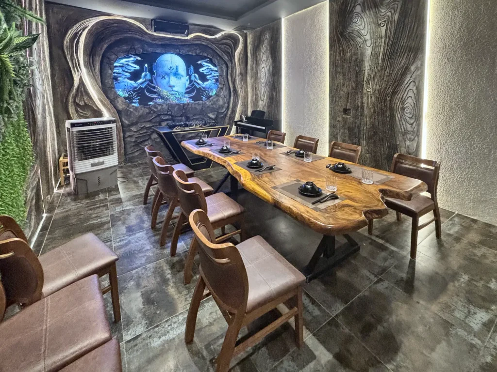 Phòng VIP 02 (Thiên Châu) tại Gánh Ngon: bàn gỗ dài, ghế bọc nệm nâu, tường vân gỗ uốn lượn, mảng cây xanh và đèn LED viền, màn hình trình chiếu cuối phòng