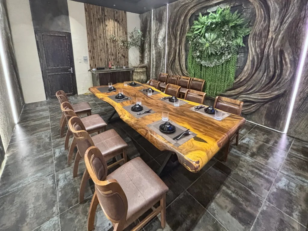 Phòng VIP 02 (Thiên Châu) tại Gánh Ngon: bàn gỗ dài, ghế bọc nệm nâu, tường vân gỗ uốn lượn, mảng cây xanh và đèn LED viền — nhìn về phía cửa ra vào