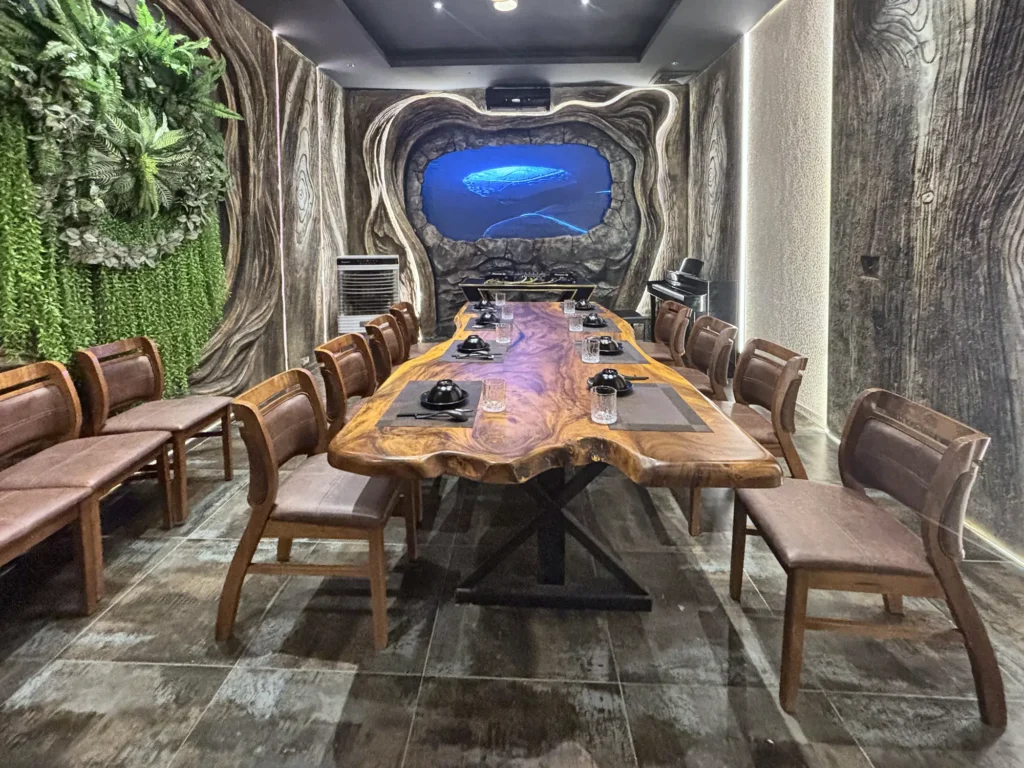 Phòng VIP 02 (Thiên Châu) tại Gánh Ngon: bàn gỗ dài, ghế bọc nệm nâu, tường vân gỗ uốn lượn, mảng cây xanh và đèn LED viền, màn hình trình chiếu cuối phòng